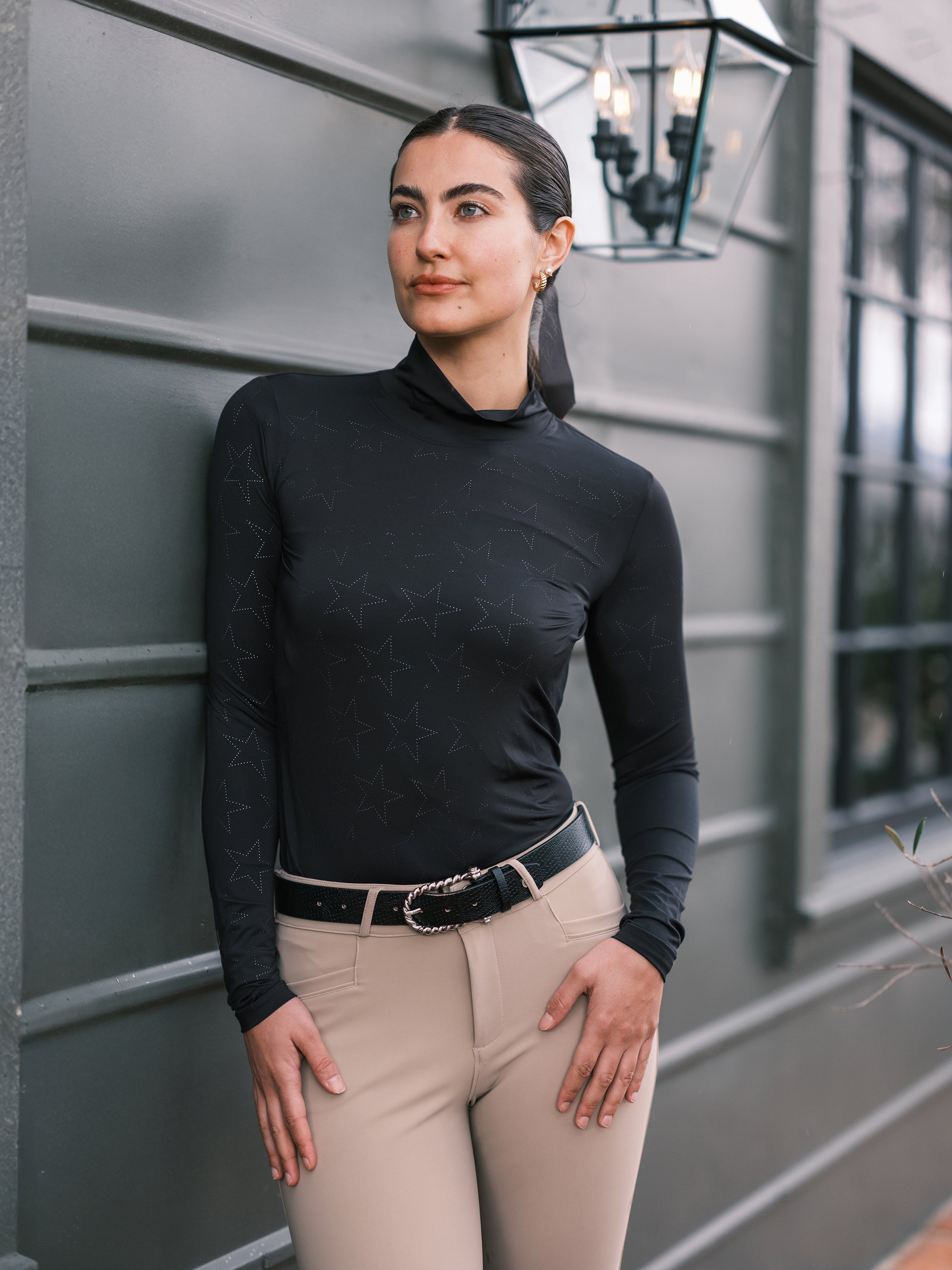 Woman wearing a black turtleneck and beige pants standing against a gray wall.